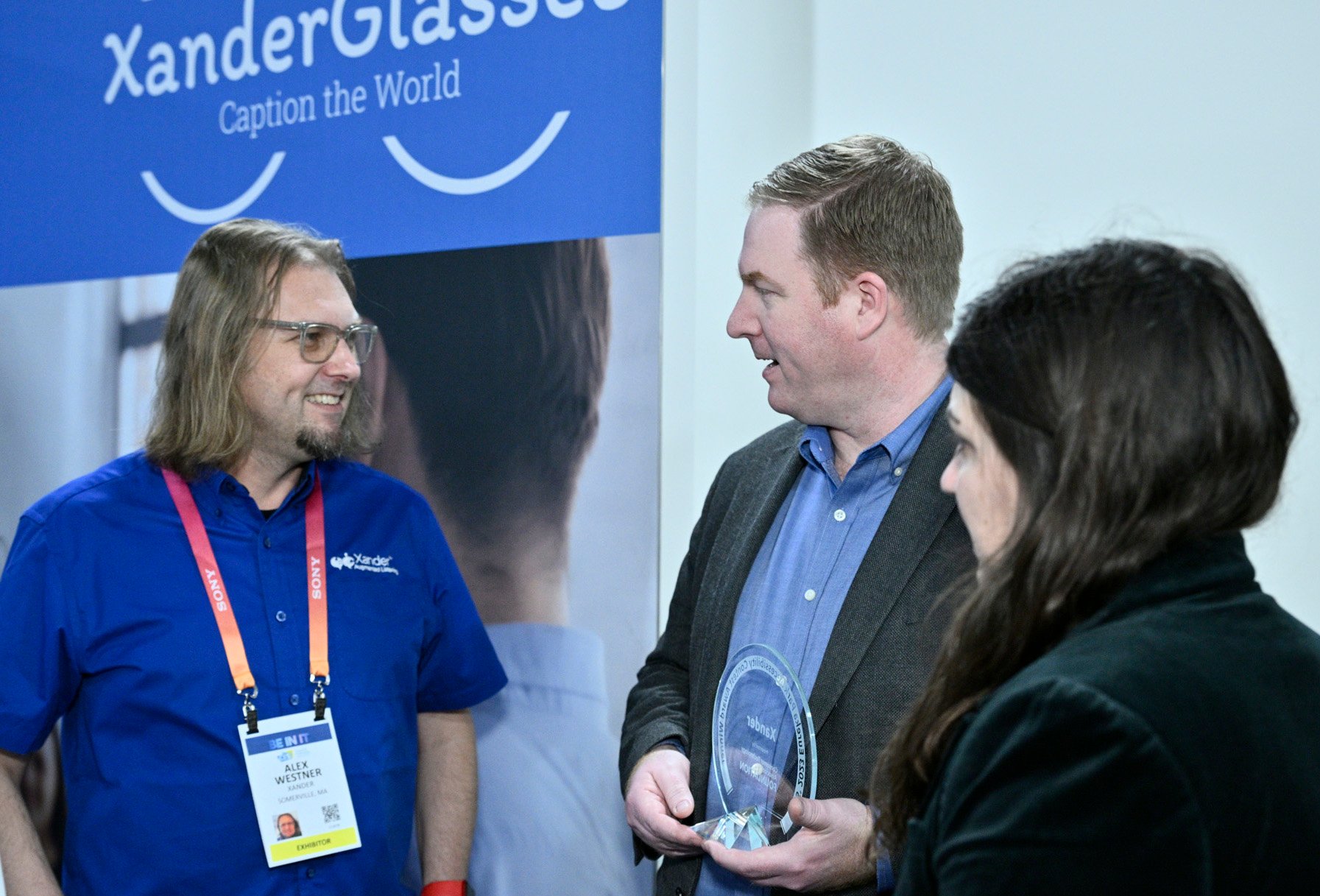 Two men and a woman standing. Man holding award to give to other man. 